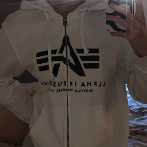 Vit hoodie från Alpha Industries - Säljer en snygg vit hoodie från Alpha Industries med svart tryck på framsidan. Hoodien har en dragkedja och justerbar huva med snören.