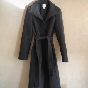 Svart Trenchcoat från H&M - Elegant svart kappa från H&M med knytbälte i midjan. Den har en stilren design med långa ärmar och en klassisk krage. Perfekt för kyligare dagar.