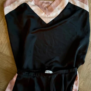 Svart och rosa linne och shorts från Hunkumöller/Denizens - Snyggt set med svart linne och matchande shorts från Denizens. Linnet har smala axelband och en v-ringad design med rosa och vita detaljer. Shortsen har elastisk midja och rosa sidopaneler. Perfekt för en avslappnad dag eller mysig hemmakväll.