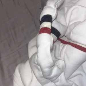 Vit hoodie med randiga detaljer - Säljer en stilren vit hoodie från Moncler med randiga detaljer i rött och blått på ärmsluten. Tröjan har en dragkedja framtill och en broderad logga på bröstet. Perfekt för en casual look!(nfc Scan finns)