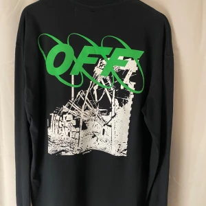 Svart långärmad tröja från Off-White - Säljer en svart långärmad tröja från Off-White med tryck både fram och bak. Framsidan har texten 'Fall-winter 19-20' och ett grafiskt motiv. Baksidan pryds av ett stort grönt 'OFF' med ett liknande motiv. Perfekt för dig som gillar streetwear!