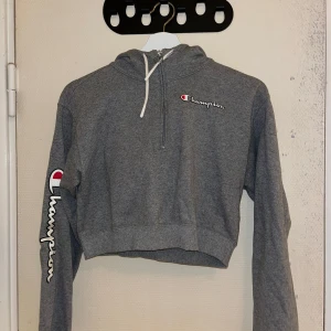 Grå croppad hoodie från Champion - Säljer en grå croppad hoodie från Champion med dragkedja och justerbar huva. Den har Champion-loggan på bröstet och ärmen. Perfekt för en avslappnad stil eller till träningen.