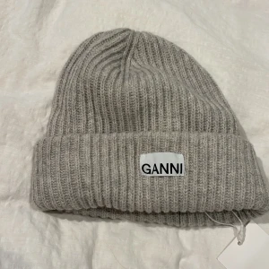Grå ribbad mössa från Ganni - Snygg och stilren grå ribbad mössa från Ganni. Perfekt för kyliga dagar med sin mjuka och bekväma passform. Mössan har en uppvikt kant och ett diskret Ganni-märke framtill. Aldrig använd med prislappen kvar