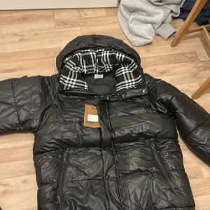 Svart dunjacka från Burberry - Snygg svart dunjacka från Burberry med klassiskt rutigt foder. Jackan har dragkedja och knappar framtill samt en hög krage för extra värme. Perfekt för kyliga dagar. Köpt för 1200 och den är endast provad så tagen är kvar .storlek L men motsvara M 