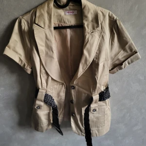 Beige kortärmad kavaj från Orsay - Snygg beige kavaj från Orsay med korta ärmar och ett flätat bälte i midjan. Kavajen har knappar framtill och två fickor. Perfekt för en stilren look.