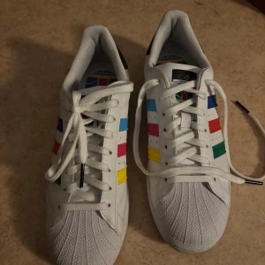 Adidas Superstar inclusivity - Säljer ett par vita Adidas Superstar sneakers med ikoniska färger. Skorna har snörning och den klassiska skalformade tån. Dessutom ingår skolådan samt de andra tre färgade snörerna till skorna. Röd, blå och grön. Dessa är limited edition och går ej längre att köpa i butik