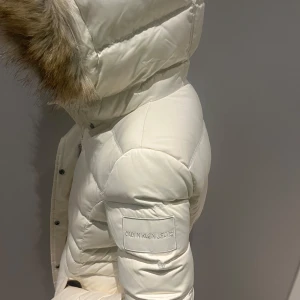 Vit dunjacka från Calvin Klein Jeans - Säljer en snygg vit dunjacka från Calvin Klein Jeans med pälskantad huva. Jackan har en quiltad design och stängs med dragkedja och knappar. Perfekt för kalla vinterdagar!