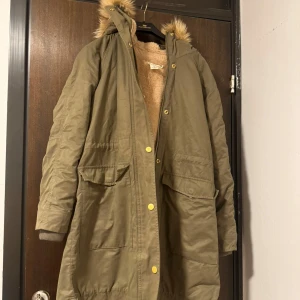 Olivgrön parkajacka med foder - Säljer en olivgrön parkajacka med mysigt foder och huva med fuskpäls. Jackan har långa ärmar och stängs med både dragkedja och knappar. Perfekt för kyliga dagar.