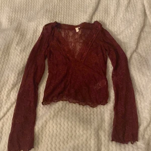 Vinröd spetsblus från Nelly - Snygg vinröd spetsblus från Nelly med omlott-design och långa ärmar. Perfekt för en elegant look med sin v-ringade stil och genomskinliga spetsdetaljer. Aldrig använd!