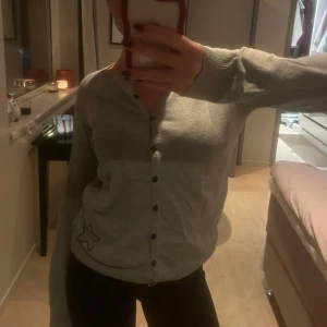 Grå kofta med stjärndetalj - Säljer en mysig grå kofta med knappar framtill och en söt stjärndetalj på sidan. Perfekt för kyliga dagar och lätt att matcha med olika outfits.