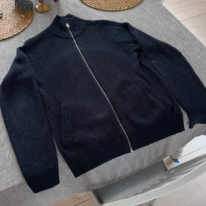 Svart stickad kofta från Jack & Jones - Säljer en stilren blå stickad kofta från Jack & Jones. Koftan har en praktisk dragkedja framtill och ribbade detaljer vid krage och ärmslut. Perfekt för kyliga dagar. Storlek M