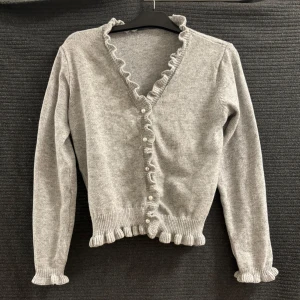 Grå kofta med volangdetaljer - Säljer en söt grå kofta med volangdetaljer längs kanterna och pärlknappar framtill. Koftan har långa ärmar och en v-ringad design, perfekt för en stilren look.