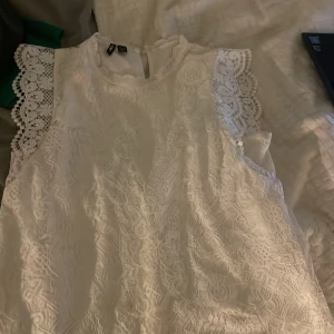 Vit spetsblus från Pieces - Säljer en vacker vit spetsblus från Pieces. Blusen är ärmlös med fina spetsdetaljer vid axlarna och har en elegant design. Perfekt för en stilren look.