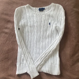 Vit stickad tröja från Ralph Lauren - Säljer en stilren vit stickad tröja från Ralph Lauren med kabelstickat mönster. Tröjan har långa ärmar och en liten broderad logotyp på bröstet. Perfekt för en klassisk look.