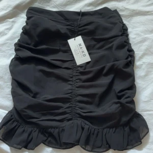 Svart kjol med volang från NA-KD - Trendig svart kjol från NA-KD med volangdetaljer. Perfekt för att skapa en stilfull look. Kjolen har en kort längd och är tillverkad i ett mjukt material. Helt ny med prislapp kvar 