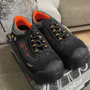 Svarta och orangea arbetsskor från Jalas - Säljer ett par svarta arbetsskor från Jalas med orangea detaljer och snörning. Skorna har en robust design med förstärkt tå och är perfekta för arbete där säkerhet är viktigt. De har en sportig look med mesh-detaljer för extra ventilation.Skorna har aldrig används.