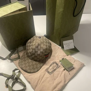 Beige keps från Gucci - Snygg beige keps från Gucci med det klassiska GG-mönstret. Kepsen har en justerbar rem baktill och en röd-grön randdetalj på sidan. Perfekt för att ge din outfit en lyxig touch. Snabb affär kan gå ned i pris. Kvitto, box, bag allt finns som ni ser på bilden!