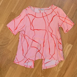 Rosa och röd topp från Minimum - Säljer en unik rosa topp från Minimum med röda abstrakta linjer. Toppen har korta ärmar och en intressant design med en slits baktill. Perfekt för att ge en färgklick till din garderob! Lite sliten i slitsen. 