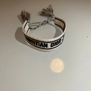Vitt armband från Christian Dior - Snyggt vitt armband från Christian Dior med svart text. Armbandet har justerbara tofsar för perfekt passform. Perfekt accessoar för att lyfta din stil.