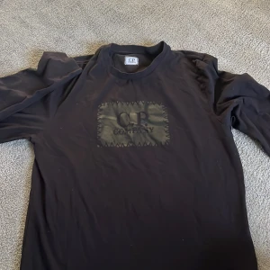 Svart långärmad tröja från C.P. Company - Snygg svart långärmad tröja från C.P. Company med ett unikt märke på bröstet. Perfekt för en stilren och avslappnad look. I storlek S.