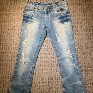 Blå jeans med slitningar - Snygga blå bootcut jeans med coola slitningar framtill. De har en klassisk femficksdesign och en unik broderad detalj på bakfickorna. Midja rakt över: 38cm. Lår: 27cm. Innerben: 78cm