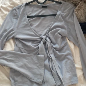 Grå långärmad topp med knyt - Snygg grå långärmad topp med v-ringning och knyt framtill. Perfekt för en stilren look. Tillverkad i mjukt material för extra komfort.
