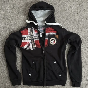 Svart hoodie från Geographical Norway - Snygg svart hoodie från Geographical Norway med tryck i rött och vitt. Den har en dragkedja framtill och en justerbar huva med vita snören. Detaljer som flaggor och text ger en cool look. Perfekt för en avslappnad stil.