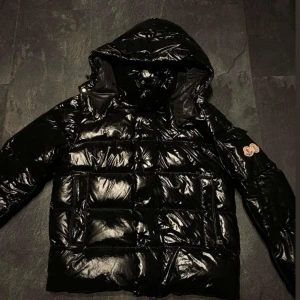 Moncler maya jacka   - Snygg svart pufferjacka med glansigt material och huva. Jackan har en quiltad design och en dragkedja framtill. Perfekt för kyliga dagar.