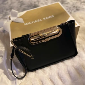 Äkta Svart axelväska från Michael Kors - Elegant svart axelväska från Michael Kors med guldfärgade detaljer och kedja. Väskan har en stilren design med ett metallhandtag och en justerbar axelrem. Perfekt för att ge en lyxig touch till din outfit.