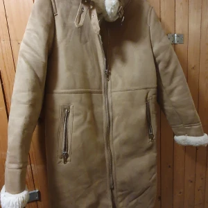 Beige pälsjacka från Zara Woman - Snygg beige pälsjacka från Zara Woman med dragkedja och bälte i kragen. Jackan har en mysig vit fuskpäls på insidan och långa ärmar. Perfekt för kyliga dagar.