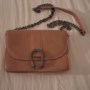 Beige axelväska från Gina Tricot - Snygg beige axelväska från Gina Tricot med kedjerem och dekorativt spänne framtill. Perfekt för att ge din outfit en stilren touch. Väskan har ett rymligt fack och stängs med magnetlås.