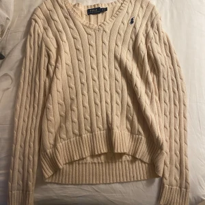 Beige stickad tröja från Ralph Lauren - Säljer en snygg beige stickad tröja från Ralph Lauren med klassiskt flätmönster och broderad logga i blått på bröstet. Tröjan har en v-ringad hals och långa ärmar, perfekt för kyligare dagar.