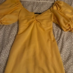 Gul klänning med puffärmar - Charmig gul klänning med puffiga kortärmar och v-ringad design. Klänningen har en snörning i ryggen som ger en fin detalj och passform. Perfekt för somriga dagar och speciella tillfällen.