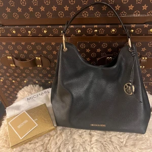 Svart axelväska från Michael Kors - Elegant svart axelväska i skinn från Michael Kors med guldfärgade detaljer och logotyp. Väskan har en rymlig insida och en praktisk axelrem. Perfekt för att bära dina dagliga nödvändigheter med stil. Väldigt bra pris för en sån fin väska, väldigt bra kvalité på den. 