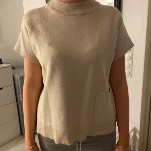 Beige stickad topp - Snygg beige stickad topp med korta ärmar och ribbad krage. Perfekt för en stilren look. Den har en praktisk ficka framtill och en avslappnad passform.