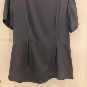 Grå blus från Ahlvar Gallery - Elegant mörkgrå blus i sidenblandning från Ahlvar Gallery. Blusen har korta ärmar och en dragkedja i ryggen för en stilren look. Perfekt för en sofistikerad stil. (Knappt använd - typ 2 ggr)