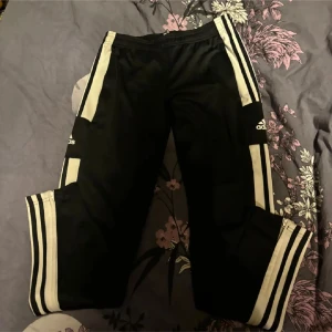 Svarta träningsbyxor från Adidas - Snygga svarta träningsbyxor från Adidas med klassiska vita ränder längs sidorna. Byxorna har en elastisk midja och är perfekta för sport och träning. Adidas-loggan syns tydligt på sidan.