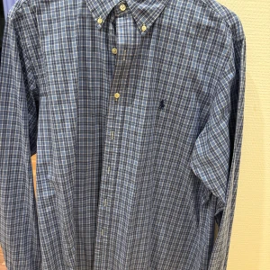 Blå rutig skjorta från Ralph Lauren - Snygg blå rutig skjorta från Ralph Lauren med klassisk krage och knappar framtill. Skjortan har långa ärmar och en broderad logotyp på bröstet. Perfekt för en stilren look.