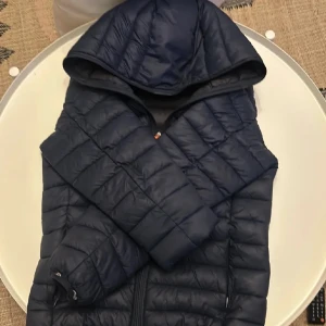 Mörkblå pufferjacka med huva - Säljer en mörkblå pufferjacka med huva. Jackan har en dragkedja framtill och är quiltad för extra värme. Perfekt för kyliga dagar.