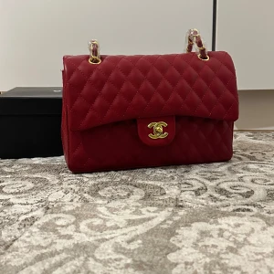 Röd quiltad axelväska  - Snygg röd axelväska  i quiltat skinn med gulddetaljer. Väskan har en klassisk design med en ikonisk logotyp på framsidan och en praktisk dragkedja inuti. Perfekt för att ge en touch av lyx till din outfit.