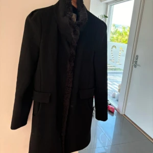 Svart kappa med pälsdetaljer - Elegant svart kappa med mjuka pälsdetaljer längs kragen och insidan. Perfekt för kyligare dagar med sin stilrena design och långa ärmar. Två fickor framtill ger extra funktionalitet.