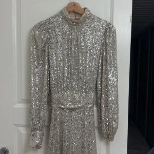 Glittrig långklänning med knytband - Snygg glittrig långklänning i silver med långa ärmar och ett elegant knytband i midjan. Perfekt för en festlig kväll med sin skimrande design.