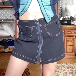 Mörkblå kjol med dragkedja - Trendig mörkblå kortkjol med framträdande dragkedja och kontrastsömmar i orange och vit. Kjolen har även små fickor fram.  Längd: 35,5cm Bredd: 35cm