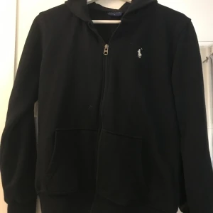 Blå hoodie från Ralph Lauren - Säljer en klassisk blå hoodie från Ralph Lauren med dragkedja och huva. Den har en liten vit logotyp på bröstet och praktiska fickor framtill. Perfekt för en stilren och bekväm look.