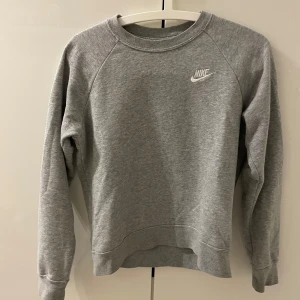 Grå sweatshirt från Nike - Säljer en klassisk grå sweatshirt från Nike med rund hals och långärmade raglanärmar. Tröjan har en broderad Nike-logga på bröstet och är tillverkad i mjukt material för extra komfort. Perfekt för en avslappnad stil.