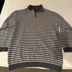 Grå randig tröja från Yves Saint Laurent - Snygg grå randig tröja från Yves Saint Laurent med dragkedja och krage. Perfekt för en stilren look. Tröjan har långa ärmar och ribbade muddar vid ärmslut och nederkant.