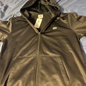 Svart hoodie från Lacoste - Snygg svart hoodie från Lacoste med dragkedja och huva. Perfekt för en casual look. Den har en klassisk krokodil-logga på bröstet och praktiska fickor framtill.