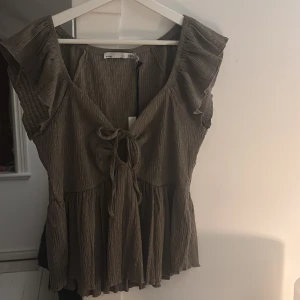 Grå topp - Säljer en söt grå blus med volangdetaljer och knyt framtill. Blusen har en v-ringad design och är ärmlös. Perfekt för fest eller fina middagar! (Passar S-M, aldrig använd)