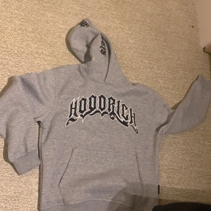 Grå hoodie från Hoodrich - Säljer en grå hoodie från Hoodrich med stort tryck på framsidan. Den har en klassisk känguruficka och en bekväm huva. Perfekt för en avslappnad stil.