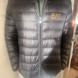 Svart lättviktsjacka från Emporio Armani - Snygg svart lätt dunjacka  från Emporio Armani med huva och dragkedja. Jackan har en quiltad design och EA7-logga på bröstet. Knappt använd. Modell 185 cm 
